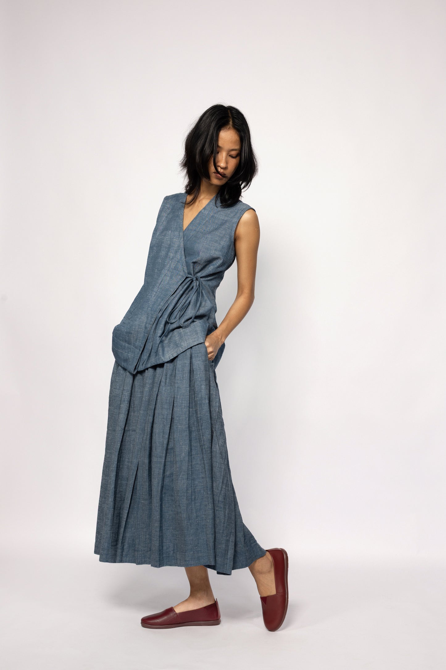 Plains Design - Lake Denim Skirt. Midi-length denim skirt with a pleated design. Paired with a matching denim sleeveless top and brown leather shoes. The denim is handmade in cotton twill.