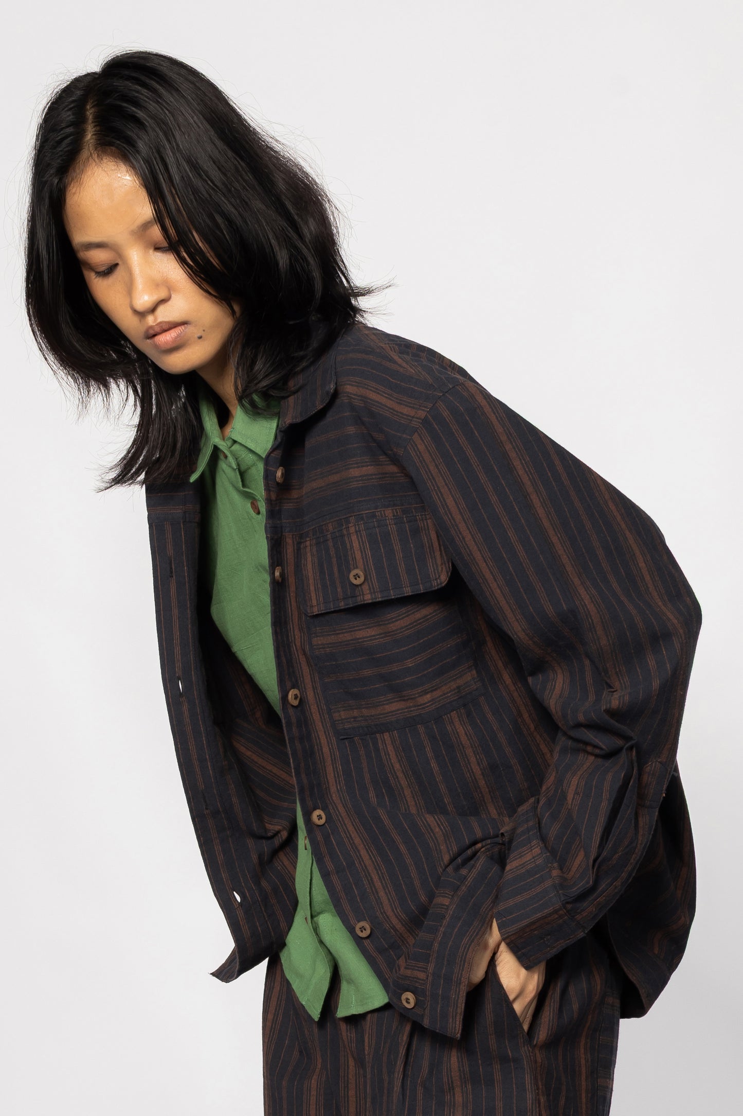 Plains Design - Brick Shirt. Oversized striped shirt in black and brick colours with long sleeves and square flap pockets on the chest. The shirt has contrasting stripes in a different direction on the hem. Worn as a jacket with a green shirt underneath.