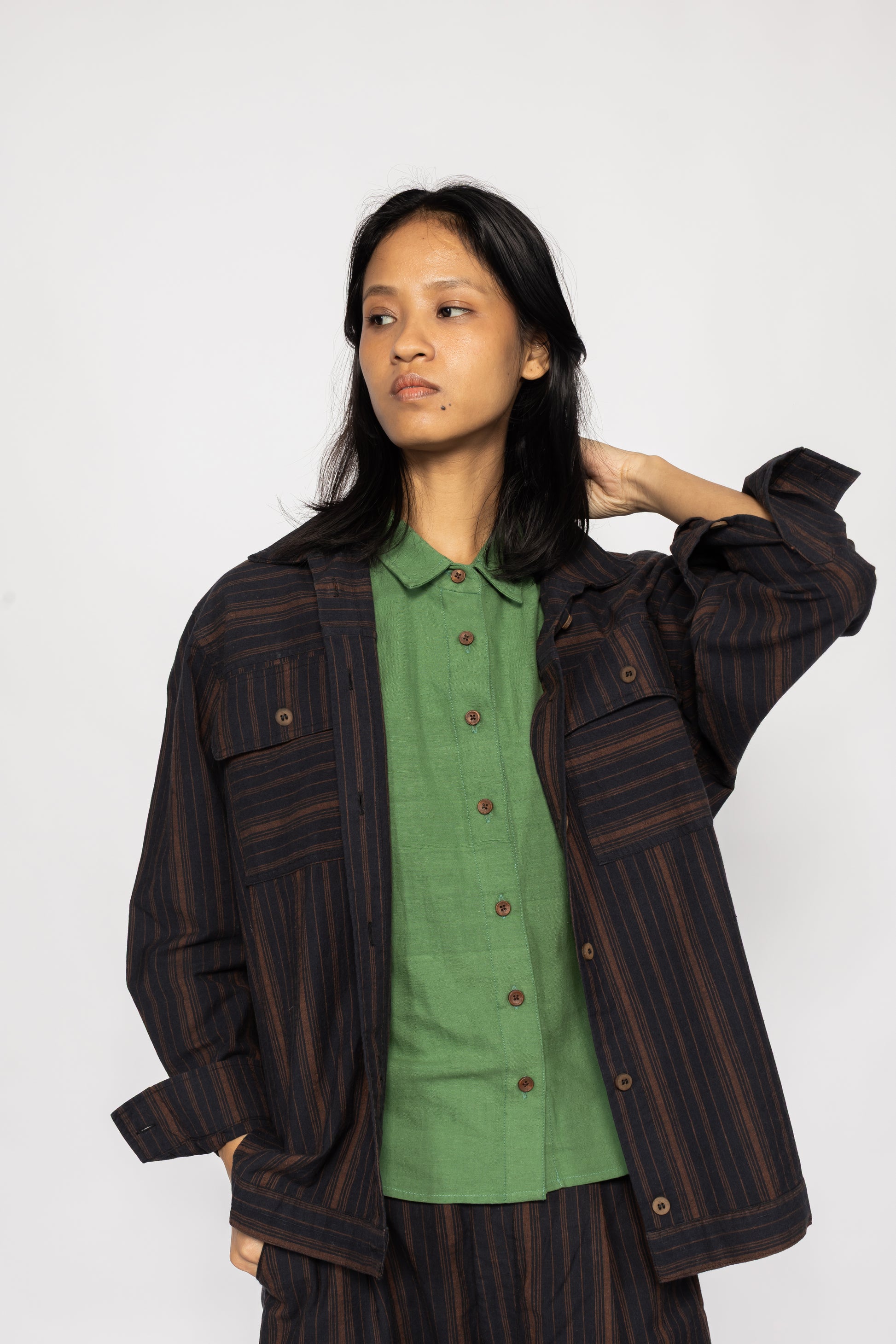Plains Design - Brick Shirt. Oversized striped shirt in black and brick colours with long sleeves and square flap pockets on the chest. The shirt has contrasting stripes in a different direction on the pockets and hem. Paired with pants of the same handwoven cotton fabric, and a green shirt.