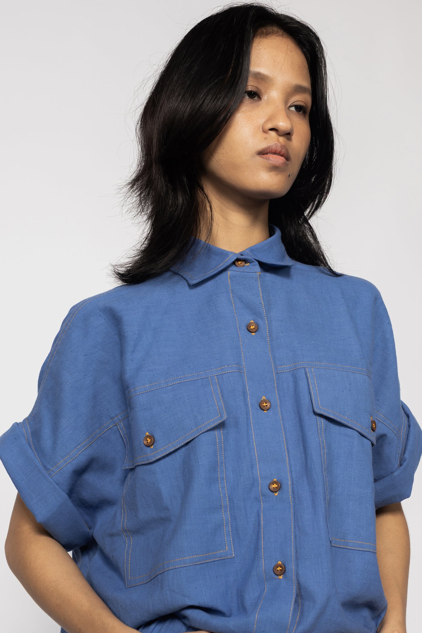 Plains Design - Trucker shirt. Front close up of a person wearing a blue cotton shirt with orange stitching, wooden buttons, short folded sleeves, and two chest pockets. Sustainable handmade cotton.