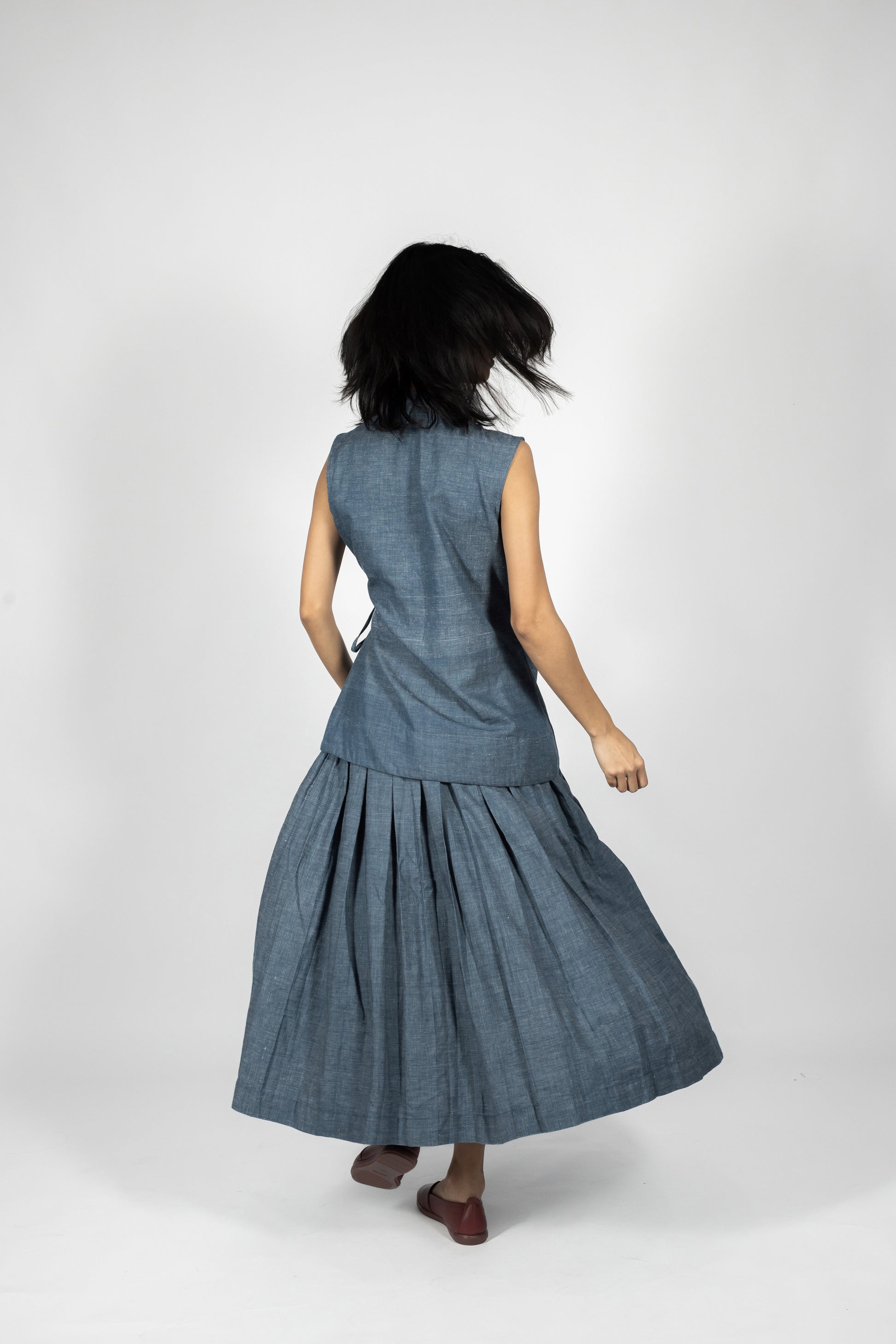 Plains Design - Lake Denim Skirt. back side of midi-length denim skirt with a pleated design. Paired with a matching denim sleeveless top and brown leather shoes. The denim is handmade in cotton twill.