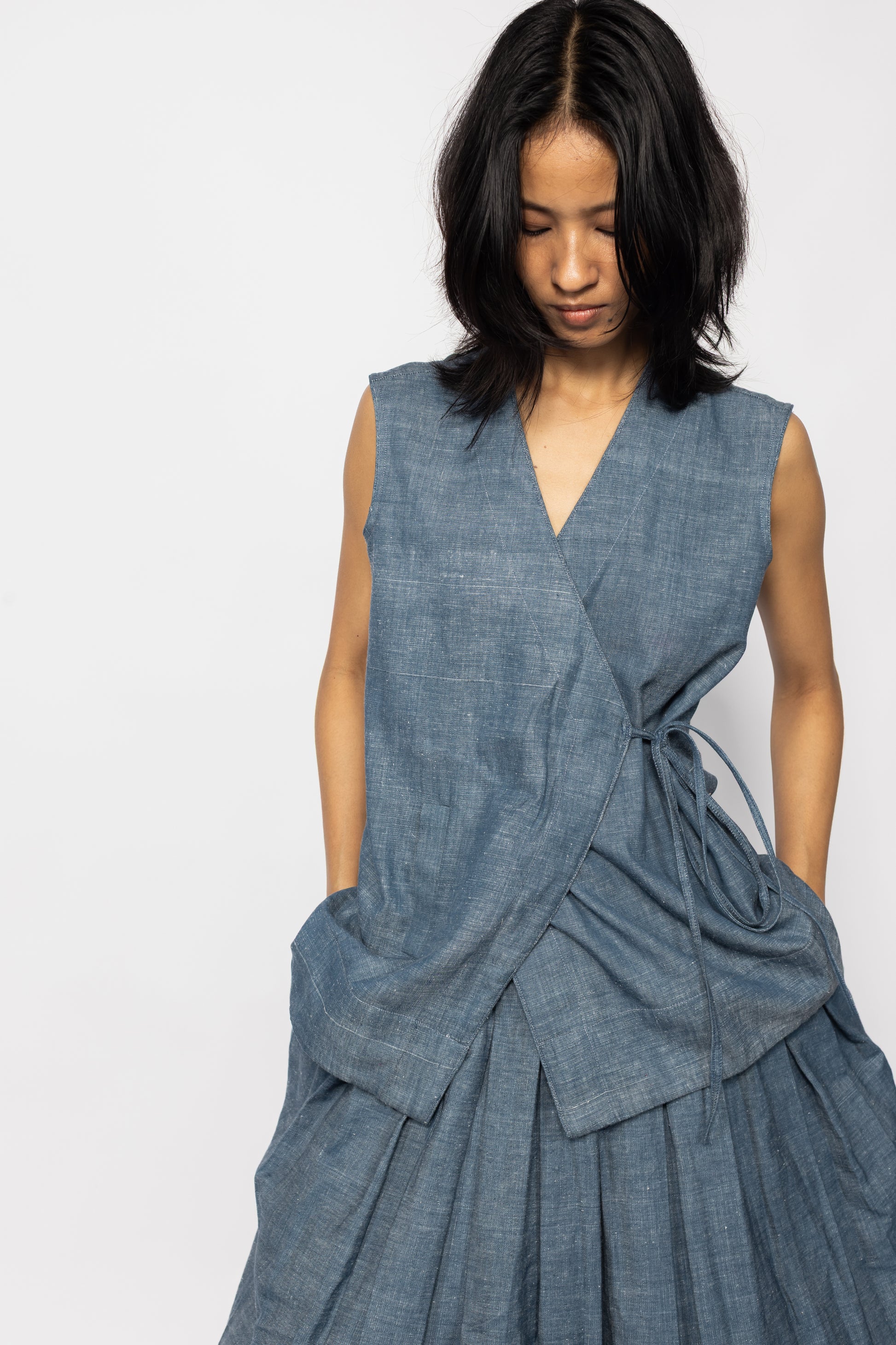 Plains Design - Wave Denim Wrap Top. Blue denim wrap top with a V-neckline and a slanted pocket on the front flap. Drawstring on the side. The denim is ethically handmade in cotton twill.