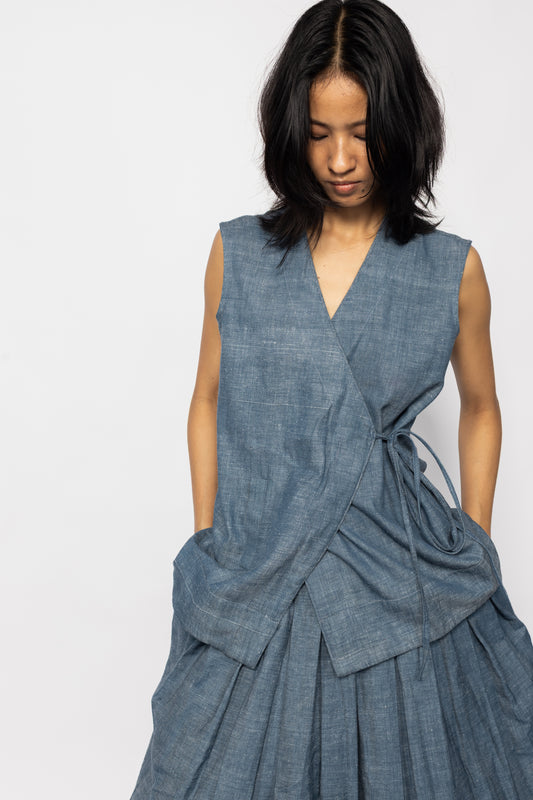 Plains Design - Wave Denim Wrap Top. Blue denim wrap top with a V-neckline and a slanted pocket on the front flap. Drawstring on the side. The denim is ethically handmade in cotton twill.