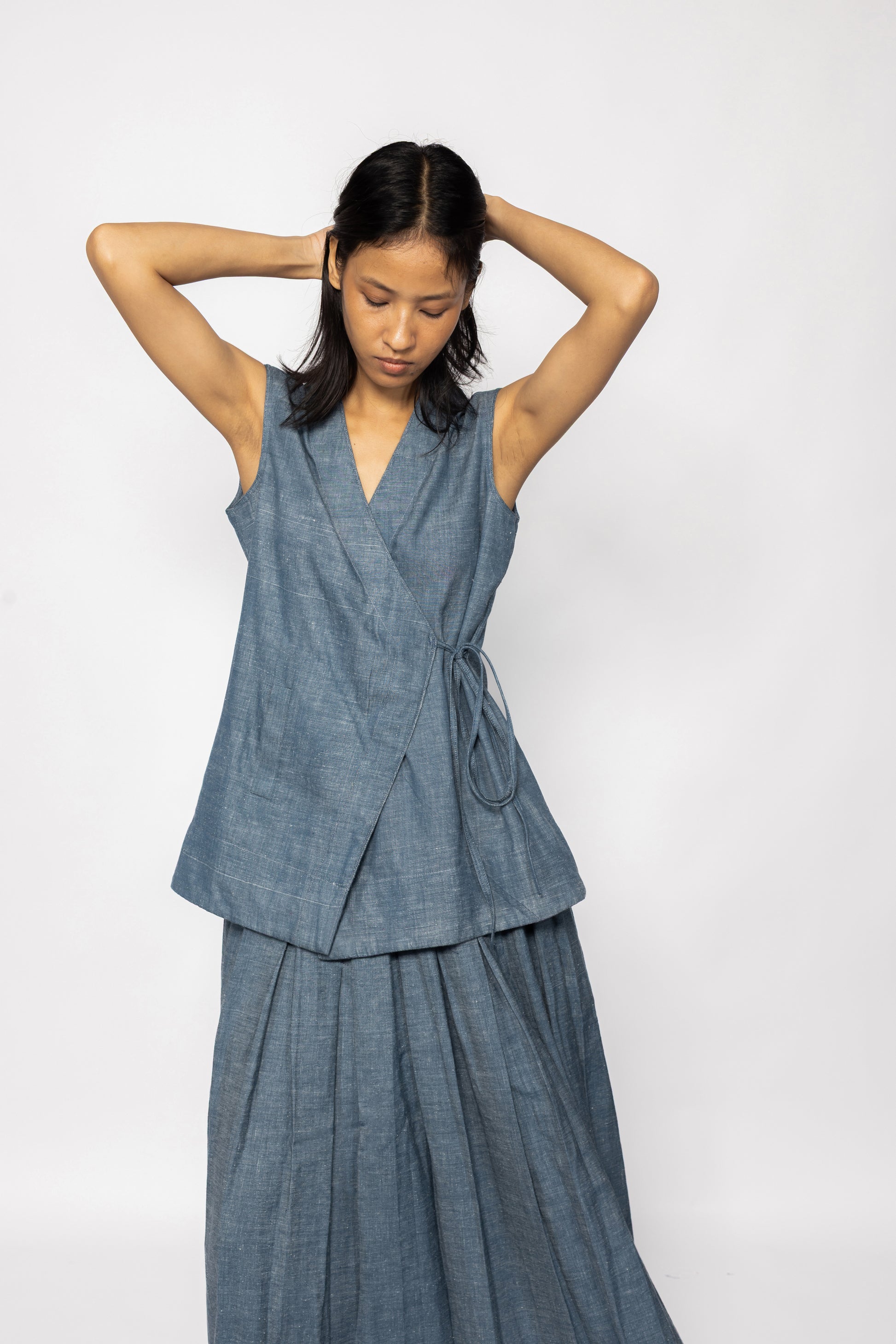 Plains Design - Wave Denim Wrap Top. Blue denim sleeveless wrap top with a V-neckline and a slanted pocket on the front flap. Drawstring on the side. Paired with a skirt of the same fabric. The denim is ethically handmade in cotton twill.