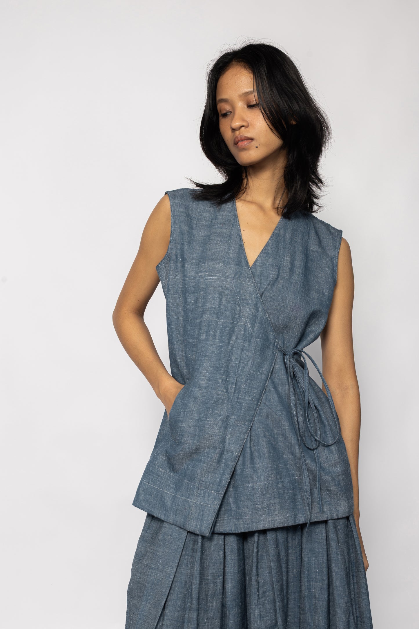 Plains Design - Wave Denim Wrap Top. Blue denim sleeveless wrap top with a V-neckline and a slanted pocket on the front flap. Drawstring on the side. Paired with a skirt of the same fabric. The denim is ethically handmade in cotton twill.