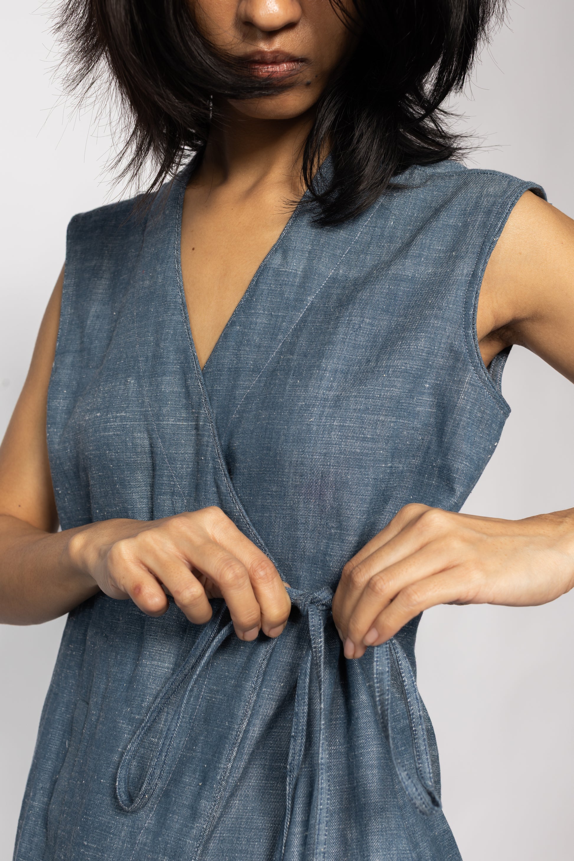 Plains Design - Wave Denim Wrap Top. Front closeup of blue denim sleeveless wrap top with a V-neckline and a slanted pocket on the front flap. Drawstring on the side. The denim is made of cotton twill. Ethically handmade in India.
