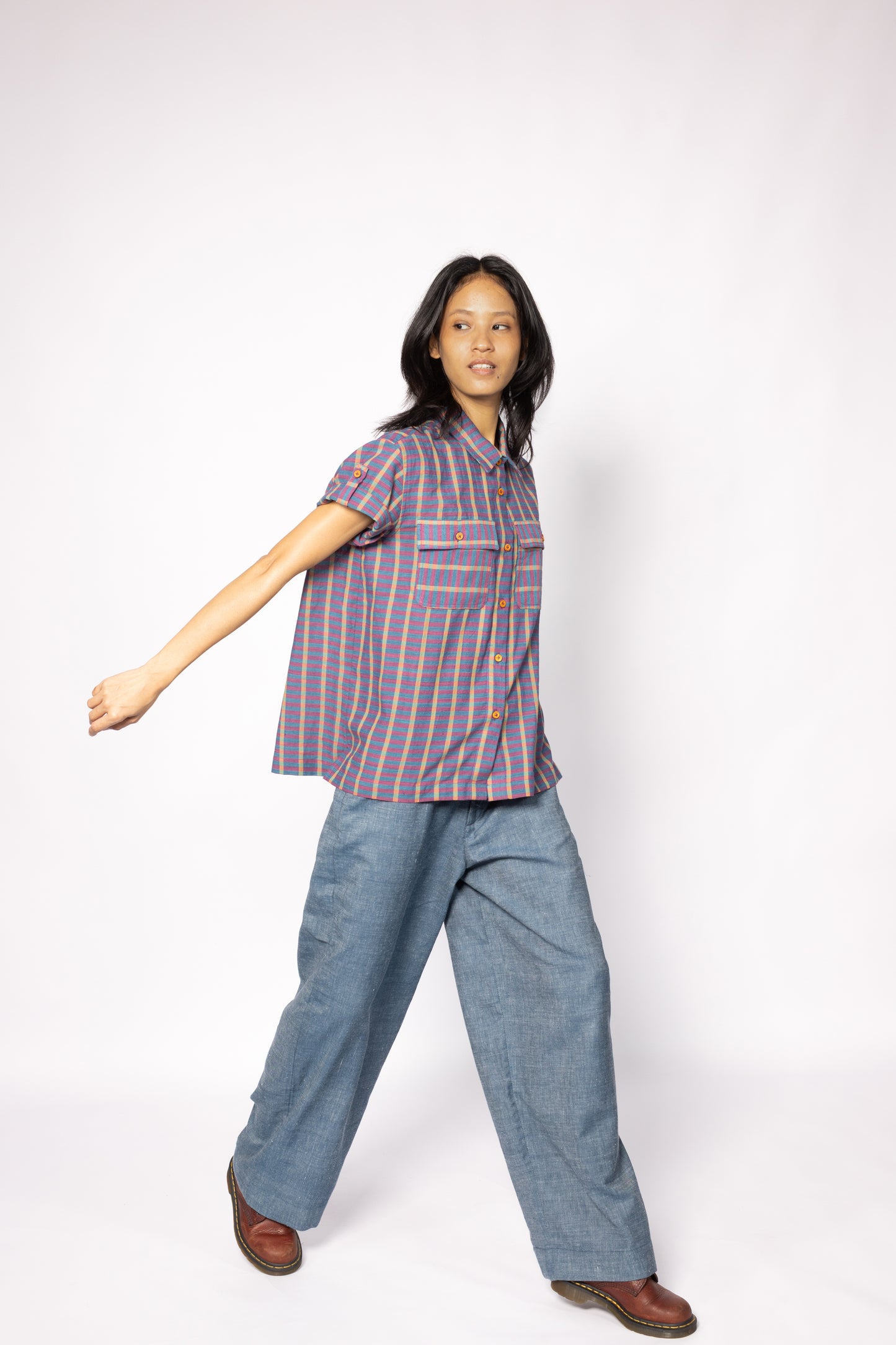 Plains Design - Twin Shirt in Purple. Striped purple and blue shirt with button-up front, short sleeves, chest pockets, and collar. Short foldable sleeves with buttons. Paired with blue denim pants and leather boots. Handmade cotton. Sustainable fashion.