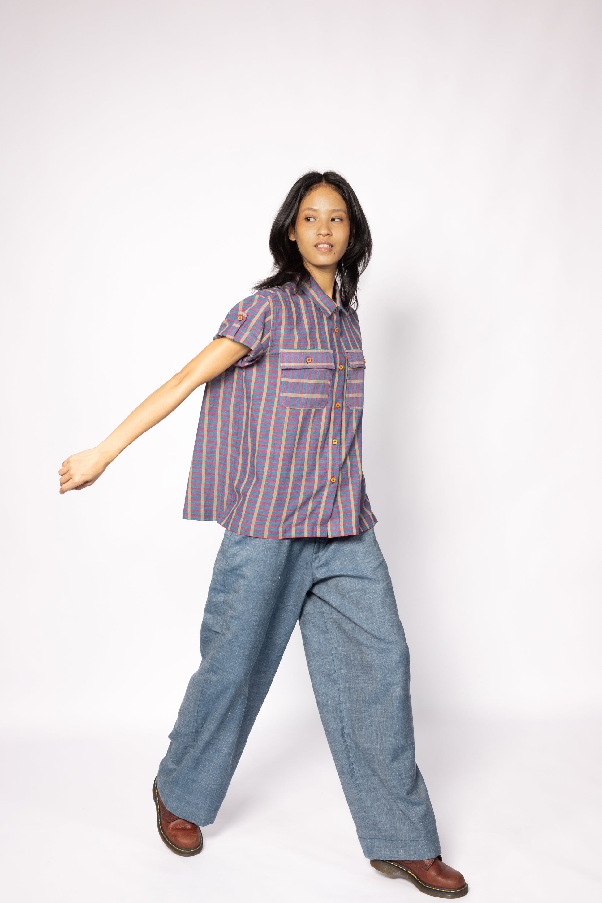 Plains Design - Twin Shirt in Purple. Striped purple and blue shirt with button-up front, short sleeves, chest pockets, and collar. Short foldable sleeves with buttons. Paired with blue denim pants and leather boots. Handmade cotton. Sustainable fashion.